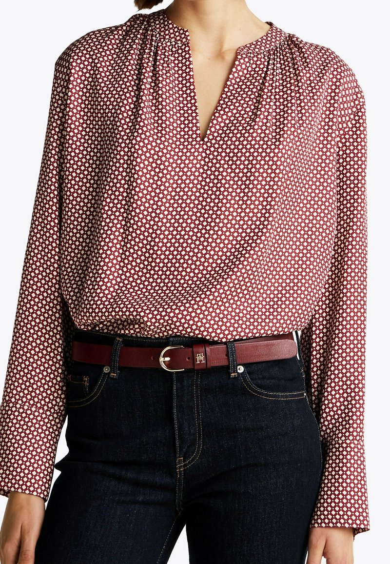 Femme portant un chemisier à manches longues rouge et blanc à motifs, rentré dans un jean foncé avec une ceinture en cuir rouge avec une boucle dorée.