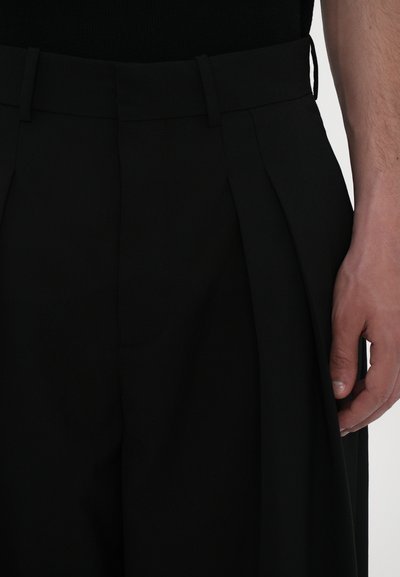 Black tailored trousers featuring a pleated front, smooth texture, and a fitted waistband, shown with a hand resting on the fabric.