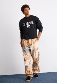 Hanorac negru cu textul "CHAMPION", asortat cu pantaloni cargo bej cu un model de camuflaj geometric și adidași negri.
