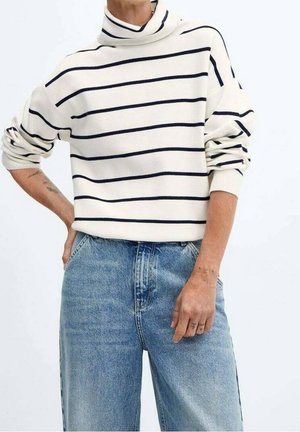 Personne portant un pull à col roulé blanc à rayures horizontales noires et un jean taille haute bleu, une main sur la hanche et l'autre détendue.