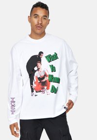 Camisa blanca de manga larga con un estampado gráfico de dos figuras y el texto "¿Qué quieres hacer?" en verde y negro. Acento púrpura en la manga.