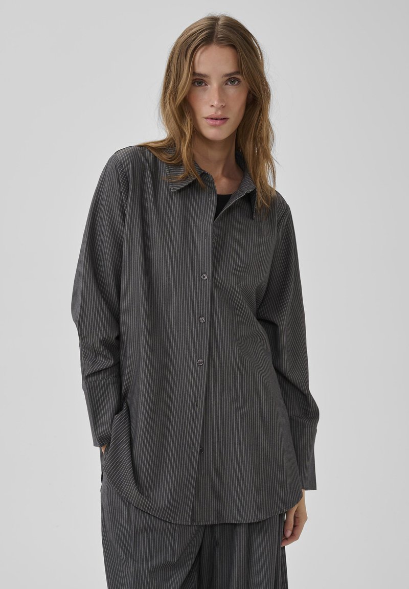 Fiatal nő, aki sötétszürke, vékony csíkos, hosszú ujjú gombos inget és hozzáillő nadrágot visel, egy egyszerű világosszürke háttér előtt áll.