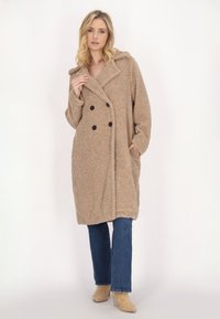 Beige, flauschiger doppelt geknöpfter Mantel mit Kapuze, ausgestattet mit schwarzen Knöpfen und seitlichen Taschen, getragen über blauen Schlagjeans und beigen Stiefeletten.