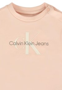 Jasnoróżowa bawełniana koszulka z okrągłym dekoltem. Zawiera teksturowane białe logo "CK" oraz napis "Calvin Klein Jeans" w ciemnoszarym kolorze.