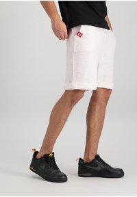 Shorts blancs texturés avec des ourlets roulés, ornés d'une étiquette de marque rouge ; associés à des chaussures Nike noires avec des accents jaunes.