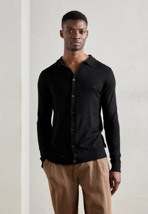 Black knit cardigan with a buttoned front, a flat collar, and ribbed cuffs. Made from soft material, worn over light brown trousers.