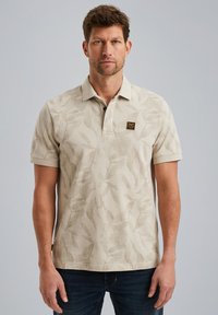 Polo shirt beige avec un motif de feuille, manches courtes et col à trois boutons. Présente un petit logo de marque noir et or sur la poitrine.