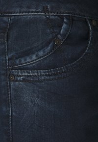 Mørkeblå denimjeans har en frontlomme med subtil søm, svart knappdetalj og en jevn tekstur.