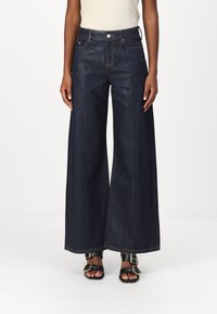 Weite dunkelblaue Jeans mit hohem Bund, zwei vorderen Taschen und Kontrastnähten. Kombiniert mit schwarzen Schnallen-Sandalen.
