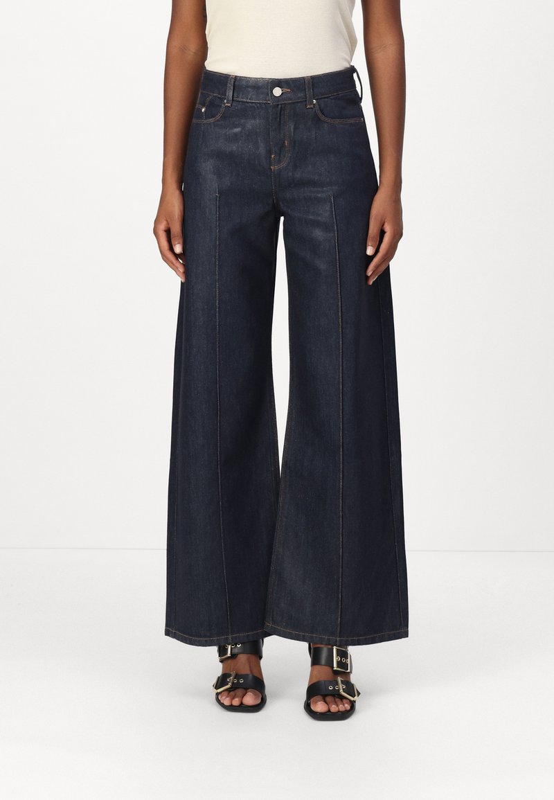Weite dunkelblaue Jeans mit hohem Bund, zwei vorderen Taschen und Kontrastnähten. Kombiniert mit schwarzen Schnallen-Sandalen.