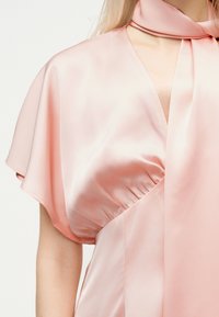 Γυναίκα που φοράει ανοιχτό ροζ σατέν φόρεμα με φτερωτά μανίκια και ταιριαστό σατέν μαντήλι στον λαιμό, δεμένο με χαλαρό κόμπο.