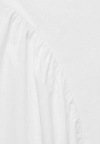Gros plan d'un vêtement blanc montrant un détail de tissu froncé le long d'une couture courbe sur un fond blanc lisse.