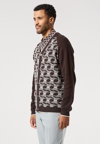 Hnědý svetr s bílým vzorovaným designem, s knoflíkovým zapínáním a žebrovanými manžetami. Přes potištěnou košili a s lehkými kalhotami.