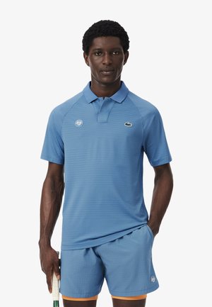 Uomo che indossa un completo da tennis blu con loghi Roland Garros, tiene una racchetta da tennis, in piedi davanti a uno sfondo semplice.