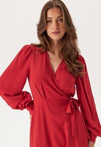 Robe rouge enveloppante avec de longues manches bouffantes, décolleté en V profond et taille nouée. Tissu lisse avec un design fluide.