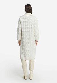 Cardigan lungo bianco a maglia con motivo a trecce, dotato di orlo e colletto a coste, abbinato a stivali al ginocchio aderenti color crema. La vista posteriore evidenzia la texture e la forma.