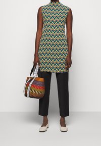 Bunt gestreiftes ärmelloses Kleid im Chevron-Muster, kombiniert mit schwarzen Hosen, einer multicolor geflochtenen Tasche und beigen flachen Schuhen.