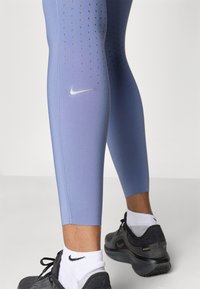 Leggings de color púrpura claro con diseño perforado, logotipo de Nike y zapatillas grises contrastantes con acentos negros y calcetines blancos.