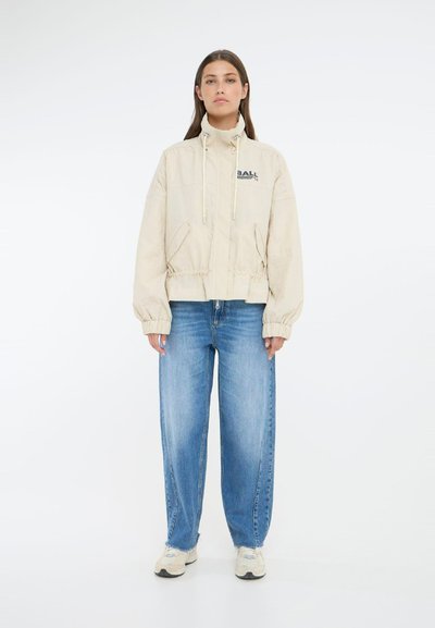 Veste beige courte avec un col montant et un cordon de serrage, arborant un logo "BALL" bleu, assortie à un jean bleu ample et des sneakers clairs.