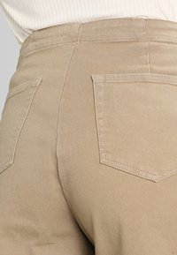 Acercamiento a unos pantalones de tiro alto beige con bolsillos traseros, combinados con una blusa blanca de canalé metida dentro.