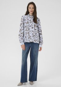 Blusa a maniche lunghe con una stampa astratta azzurro chiaro e marrone, caratterizzata da pieghe e chiusura con bottoni. Abbinata a jeans a gamba larga e scarpe beige.