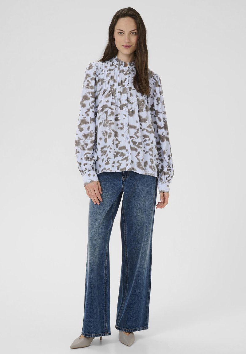 Blusa a maniche lunghe con una stampa astratta azzurro chiaro e marrone, caratterizzata da pieghe e chiusura con bottoni. Abbinata a jeans a gamba larga e scarpe beige.