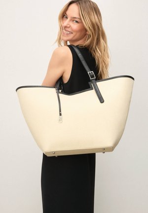 Didelė kreminės spalvos rankinė su juodos odos apvadais ir reguliuojama dirželiu; pasižymi struktūruota forma ir sidabrinės spalvos aparatūros detalėmis.