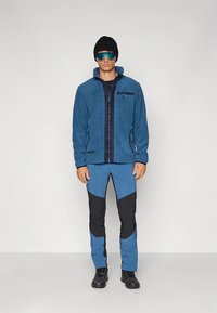 Plava flis jakna s tamnim detaljima, zatvaranjem na patentni zatvarač i bočnim džepovima, u kombinaciji s plavim i crnim hlačama koje imaju kontrastne panele i kroj prilagođen tijelu.