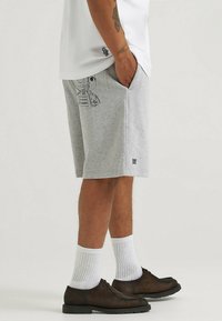 Pantaloni corti in cotone grigi con una tasca laterale che presenta un design grafico nero. Abbinati a calzini bianchi a coste e scarpe stringate marrone scuro.