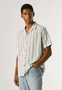 Jauns vīrietis, kurš valkā īsroku baltu kreklu ar vertikālām zilām svītrām un gaiši zilas džinsas, stāv ar vienu roku kabatā.