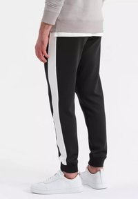 Zwarte joggerbroek met witte zijstrepen, elastische manchetten en een gladde textuur. Gedragen met witte sneakers.