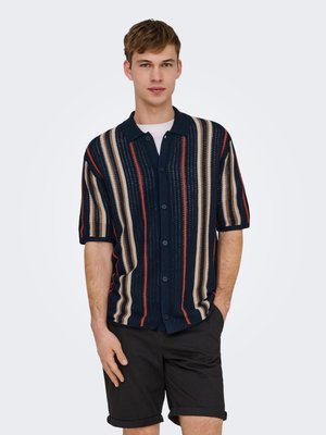 Giovane uomo in piedi con le mani in tasca, indossa una camicia a maniche corte con bottoni a righe verticali blu navy, beige e rosse e pantaloncini neri.