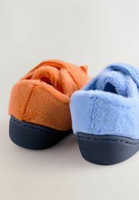 Chaussons en fausse fourrure orange et bleu, avec une forme arrondie et une semelle noire avec des détails de couture. Texture douce évidente.