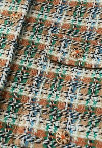 Gros plan sur un tissu tweed multicolore avec des fils verts, blancs, marron, bleus et des boutons décoratifs dorés ornés de détails noirs et orange.