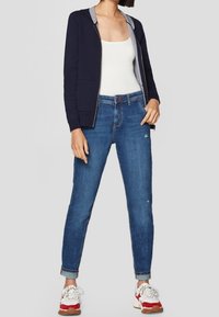 Marine blauw zip-up hoodie met grijze voering, witte tanktop, blauwe jeans met opgerolde pijpen en rood/witte sneakers met gestructureerde zolen.