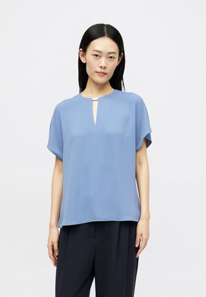 Light blue blouse with a keyhole neckline, short sleeves, and a smooth, lightweight fabric. Paired with dark trousers, presenting a relaxed silhouette.