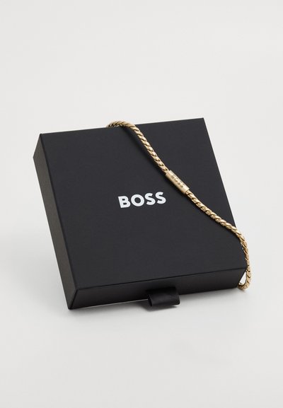 Златна плетена гривна, изложена на черна кутия с логото "BOSS" в бяло. Кутията има лента за отваряне.