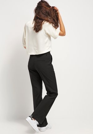 Femme aux longs cheveux bruns portant un haut blanc à manches courtes, un pantalon noir et des baskets blanches, debout dos à la caméra.