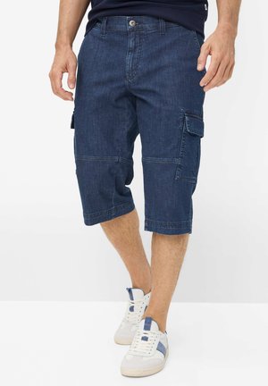 Mann trägt dunkelblaue, knielange Denim-Cargoshorts und weiße Sneakers mit blauen Akzenten, steht mit verschränkten Beinen.