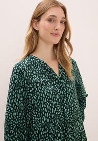 Groene blouse met een patroon, een v-halslijn, kleine knopen en losse, gerimpelde mouwen. De textuur is soepel en vertoont een speels abstract ontwerp.