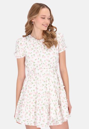 Nainen, jolla on aaltoilevat vaaleat hiukset, pukeutunut valkoiseen lyhythihaiseen mekkoon, jossa on vaaleanpunainen kirsikkakuosi ja röyhelöhelma, katsoo alaspäin ja hymyilee.