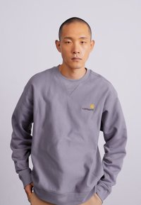 Suéter gris con cuello redondo y puños acanalados. Presenta un pequeño logo amarillo en la parte superior izquierda. Tejido suave y texturizado.