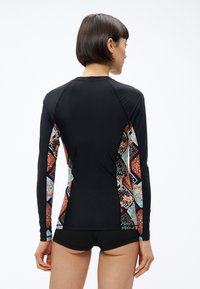 Kobieta z krótkimi ciemnymi włosami, ubrana w czarną długorękawową koszulkę do pływania z kolorowymi, wzorzystymi bokami oraz czarne szorty plażowe, stojąca tyłem.