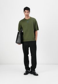 Olive green knitted short-sleeve sweater with a blue label, paired with black trousers and a black tote bag, featuring a structured design.