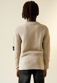 Bézs kötött pulóver bordázott mintával, kerek nyakkivágással és hosszú ujjal. A bal ujjon fekete címke található. Sötét farmerral viselve.