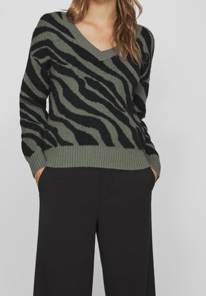 Femme portant un pull à col en V rayé zèbre vert et noir avec un pantalon noir, mains dans les poches, debout devant un fond uni.