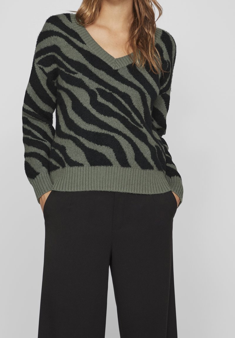 Femme portant un pull à col en V rayé zèbre vert et noir avec un pantalon noir, mains dans les poches, debout devant un fond uni.