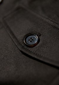 Close-up van een zwart stoffen patch met een afgeronde knop met vier gaten, gestikte details en een gladde textuur.