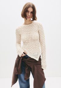 Jonge vrouw met kort bruin haar, gekleed in een crèmekleurige kanten top met lange mouwen, blauwe spijkerbroek en een bruine jas om de taille gebonden.