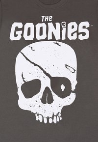 Donkergrijze T-shirt met een grote witte gebarsten schedelafbeelding en de tekst "THE GOONIES" erboven in vette letters. Zacht, katoenen materiaal.
